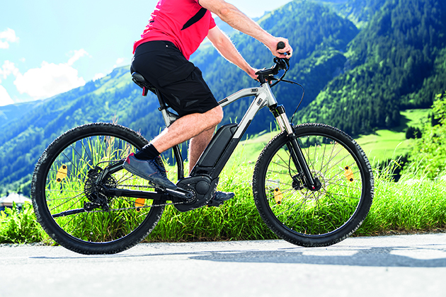 A person wearing a pink shirt and black shorts rides a mountain bike with advanced electronics on a paved road, surrounded by green grassy hills and tall mountains under a blue sky. by Standex Detect